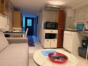 A compact living space with a sofa, a dining area with a table and chairs, a kitchen with appliances, and a hallway leading to a door.