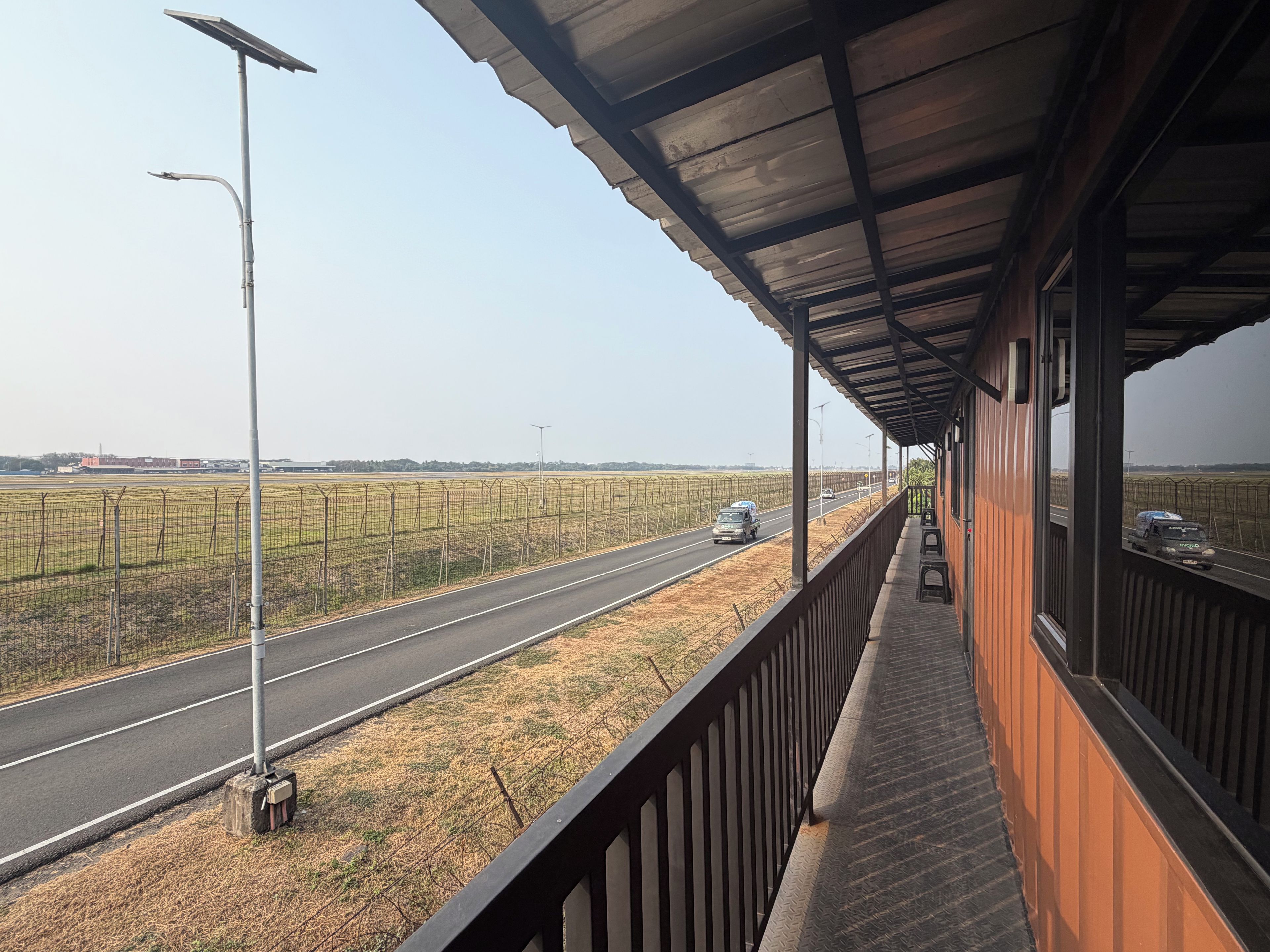 Een balkon met uitzicht op een weg, velden, een lantaarnpaal en een gebouw met een metalen dak.
