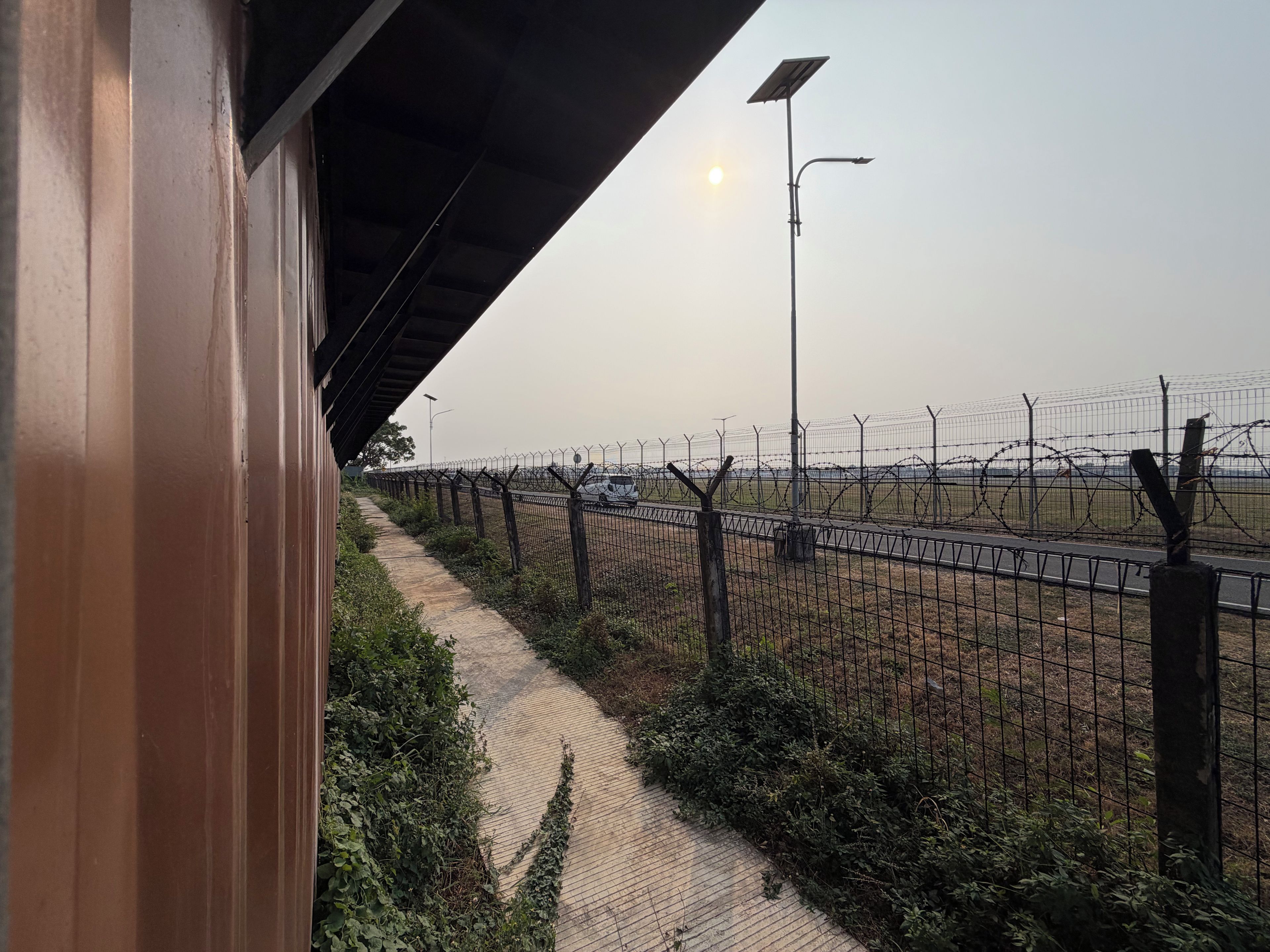 A pathway beside a fence with a streetlight and parked cars in the background.