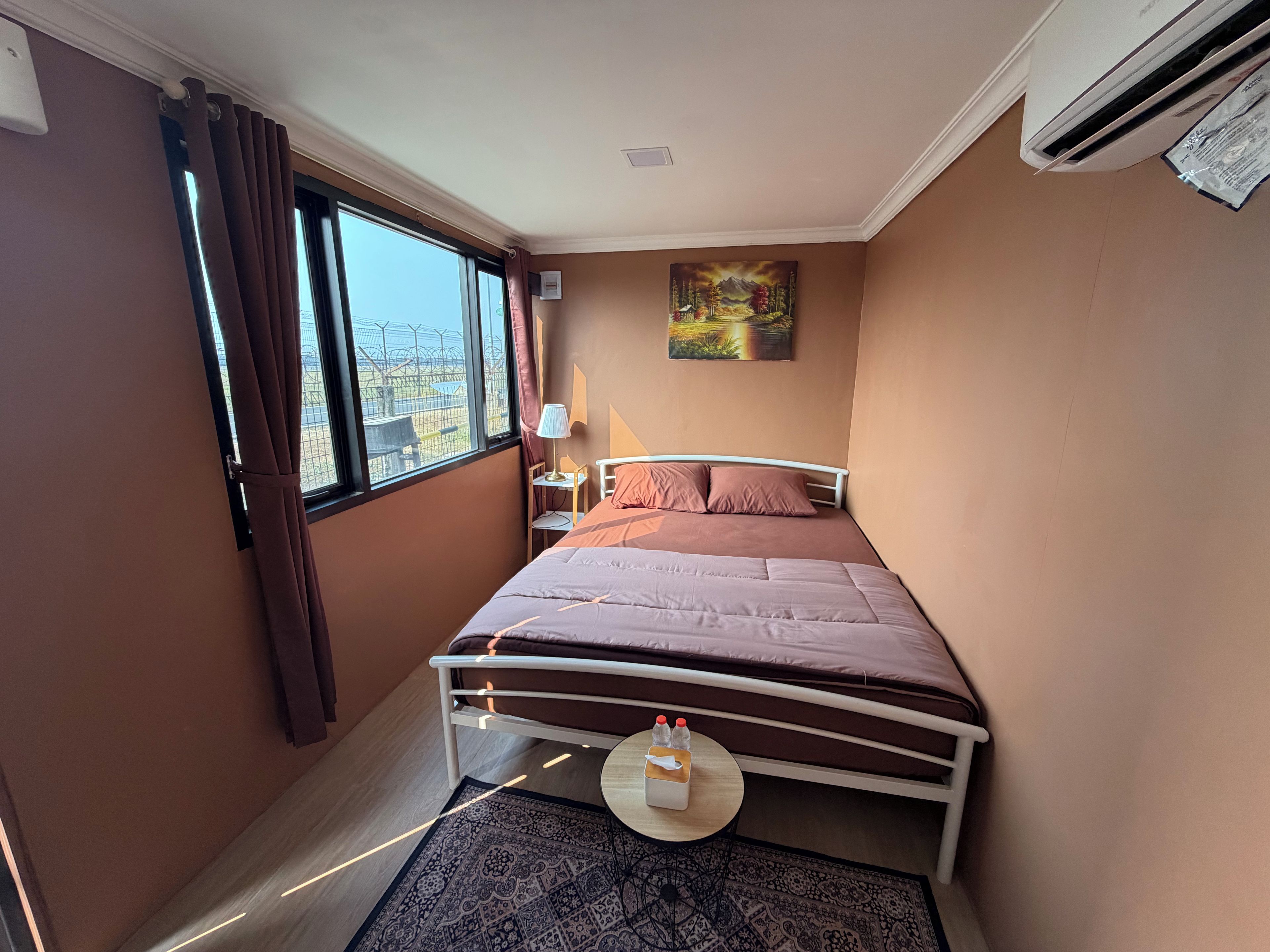 Habitación de hotel con cama, mesita de noche, lámpara, un cuadro y vistas al exterior.