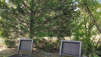 Two metal chairs with mesh backs and a glass table in a natural setting with trees and foliage.