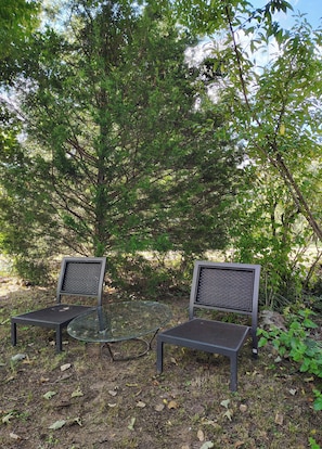 Two metal chairs with mesh backs and a glass table in a natural setting with trees and foliage.