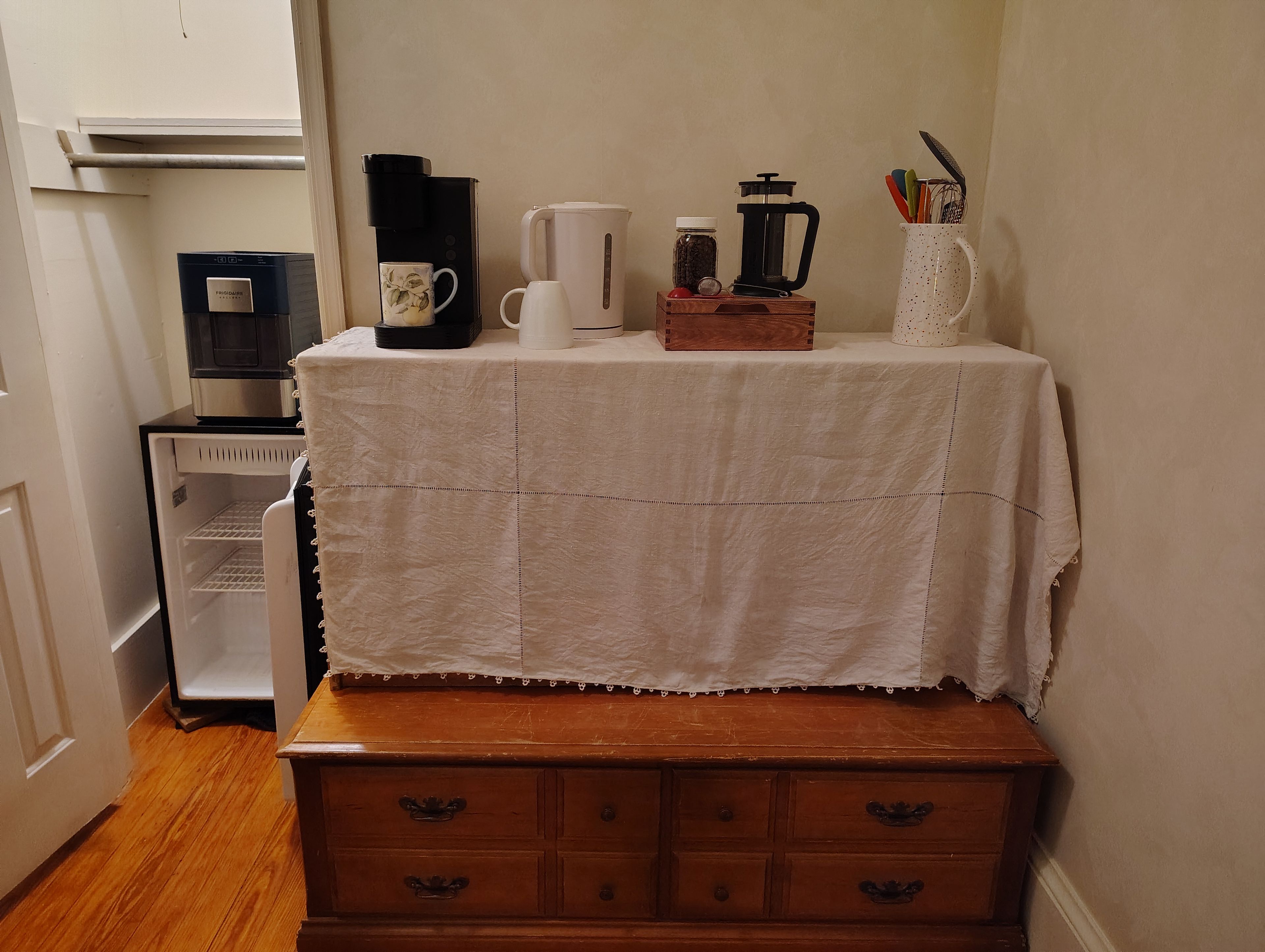A kitchenette with a coffee maker, a kettle, a jar of coffee beans, and a mug on a table.