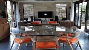 Una mesa redonda de vidrio rodeada de sillas con cojines naranjas, un televisor en la pared y vistas a zonas verdes al exterior.