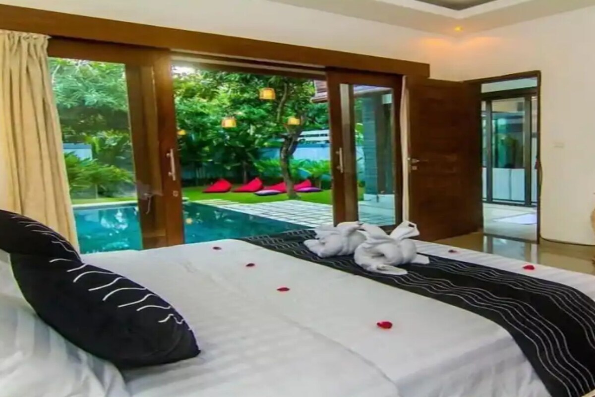 A hotel room with a bed, a desk, a TV, and a view of a pool and greenery.