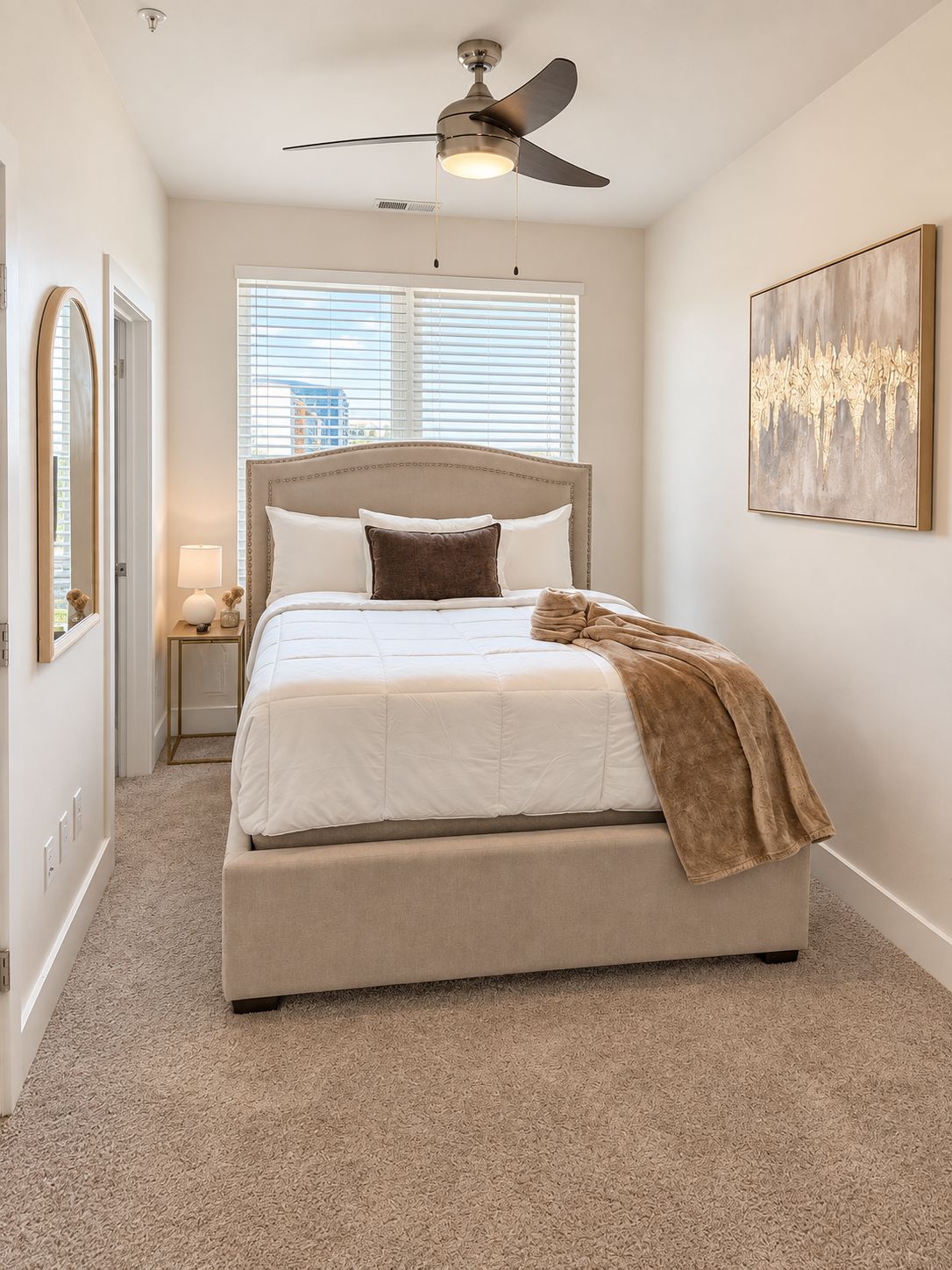 A bedroom with a bed, a ceiling fan, a window with blinds, a nightstand with a lamp, and a painting on the wall.