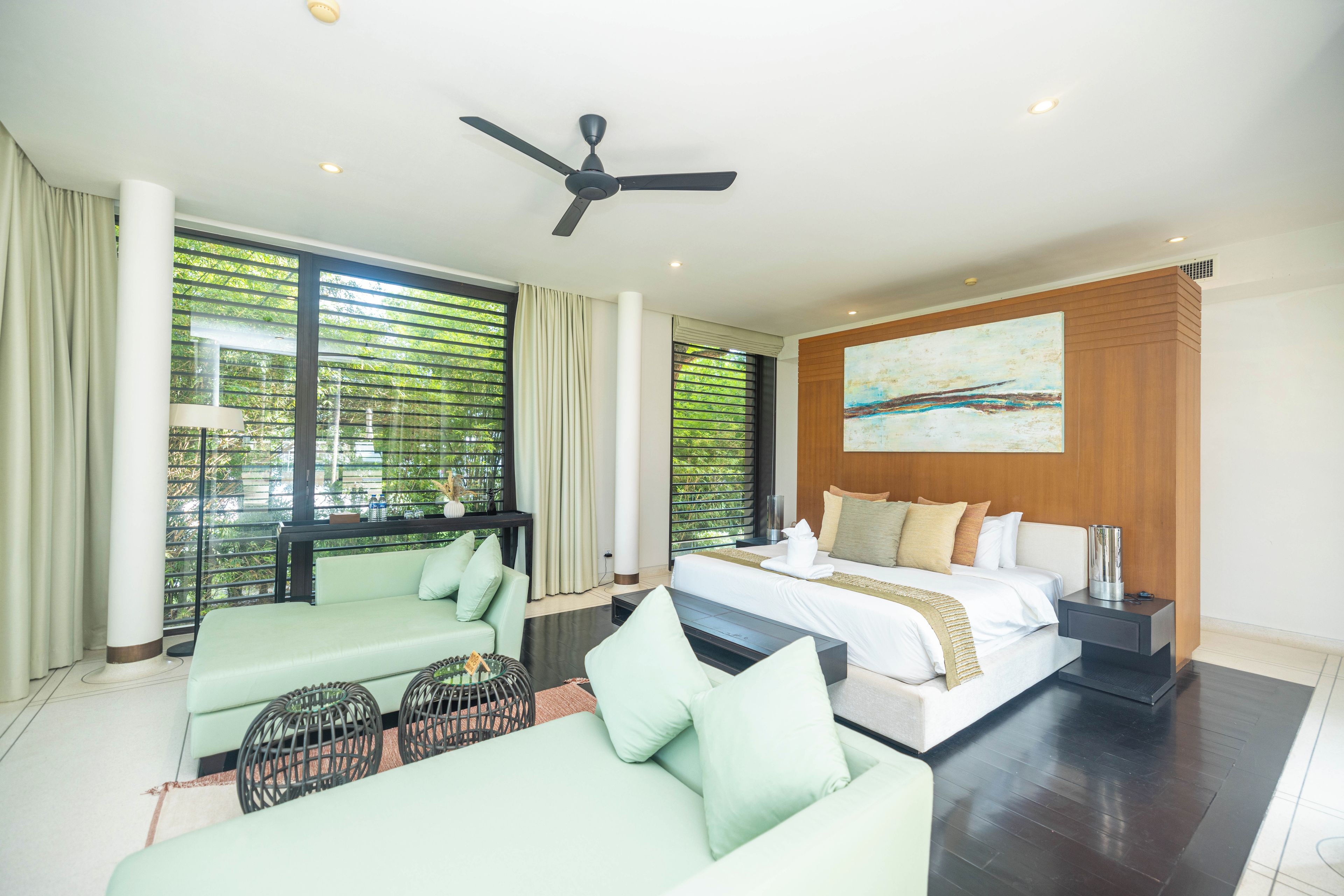 Una habitación de hotel moderna con cama, sofá, ventilador de techo y ventanas amplias con persianas.