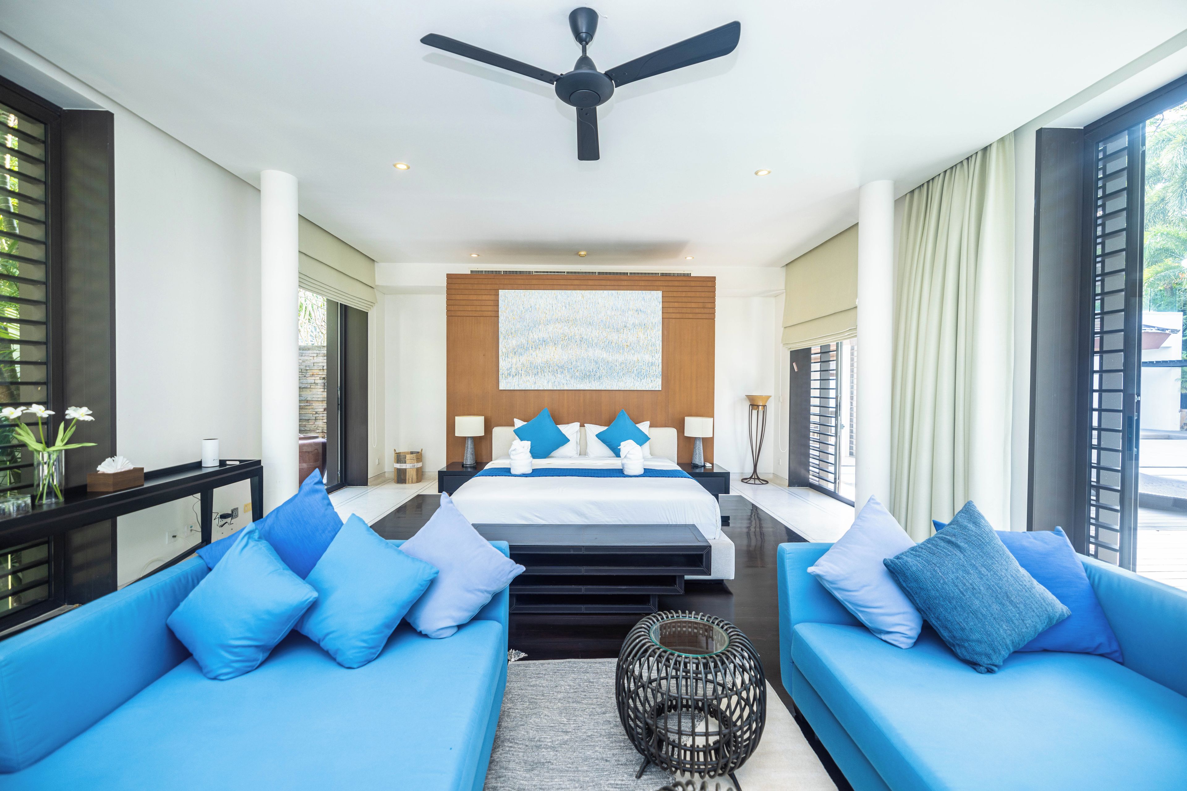 Una habitación de hotel moderna con una cama grande, dos sofás azules, un ventilador de techo y un ventanal amplio.