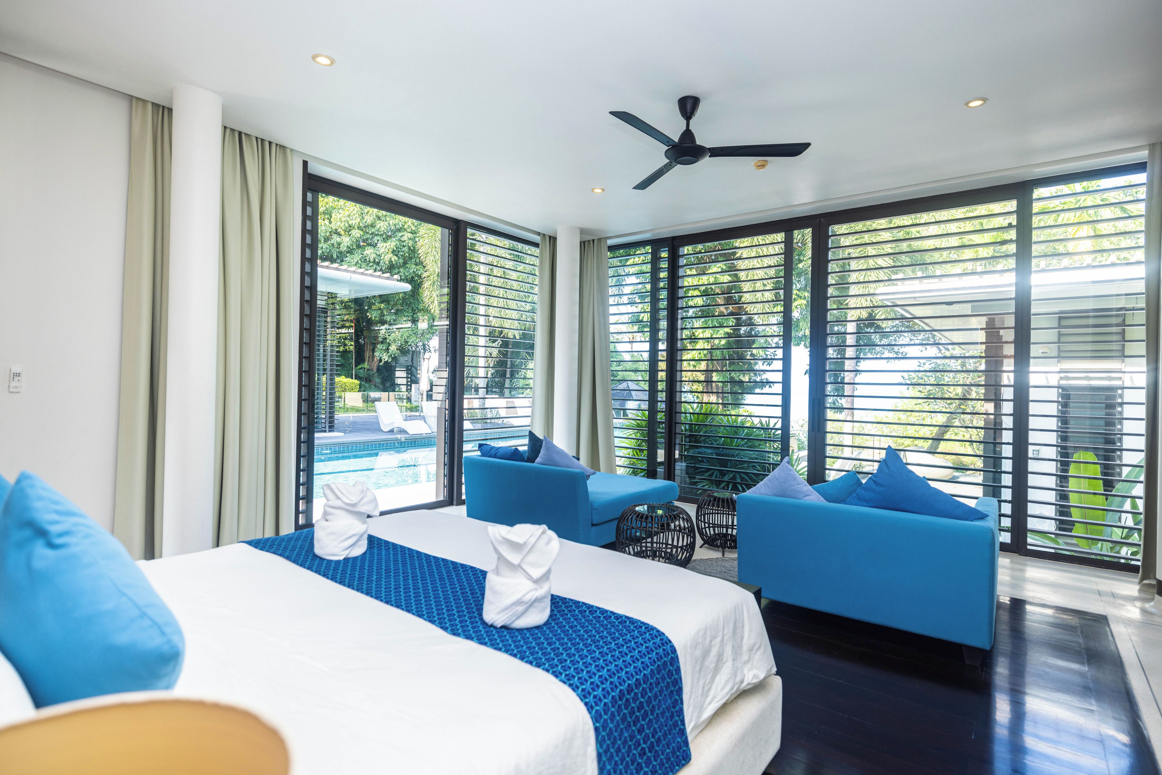 Habitación de hotel con cama, dos sofás azules, ventilador de techo y ventanas grandes con persianas.