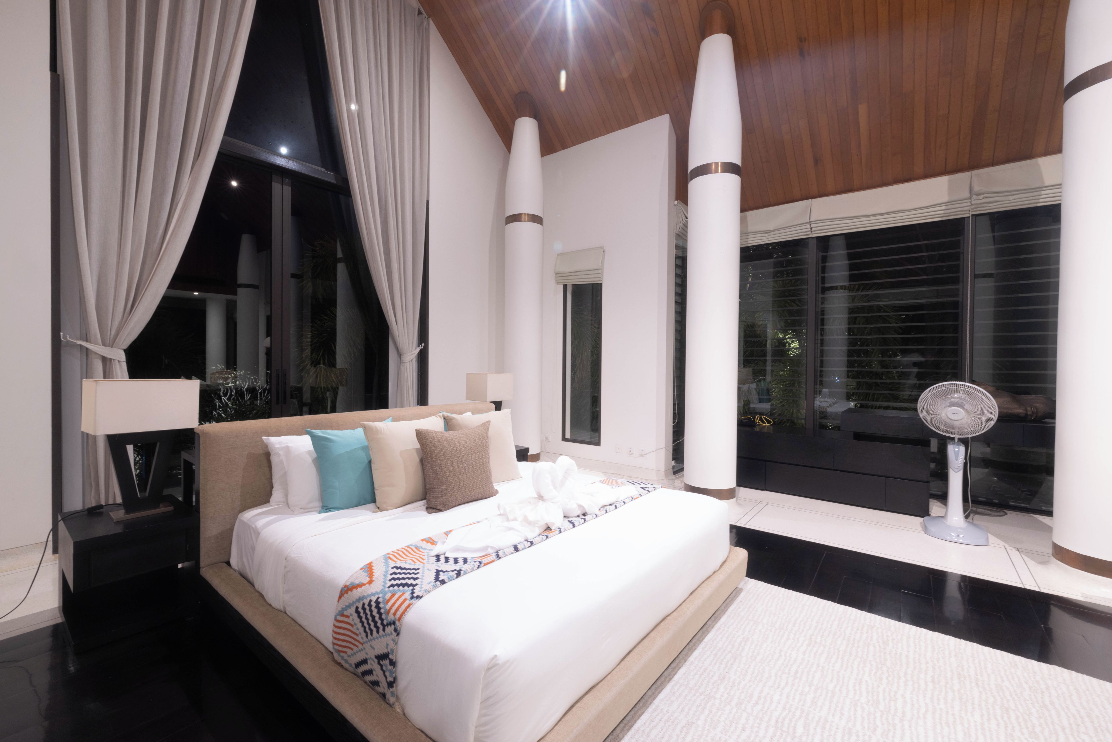 A modern hotel room with a large bed, a standing fan, and a view of the outdoors.