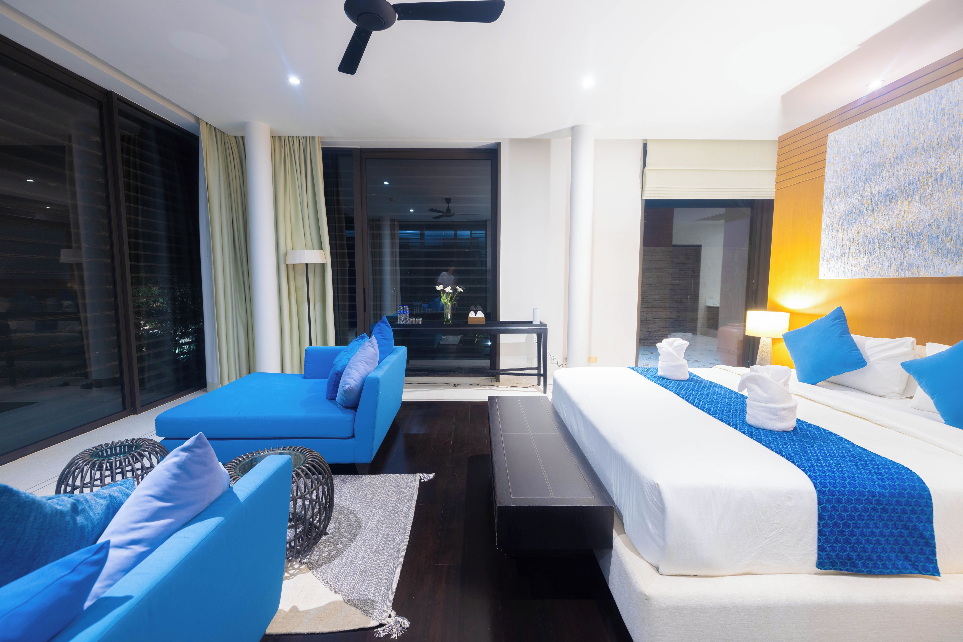 A modern hotel room with a bed, a sofa, a small table, and a view through a large window.