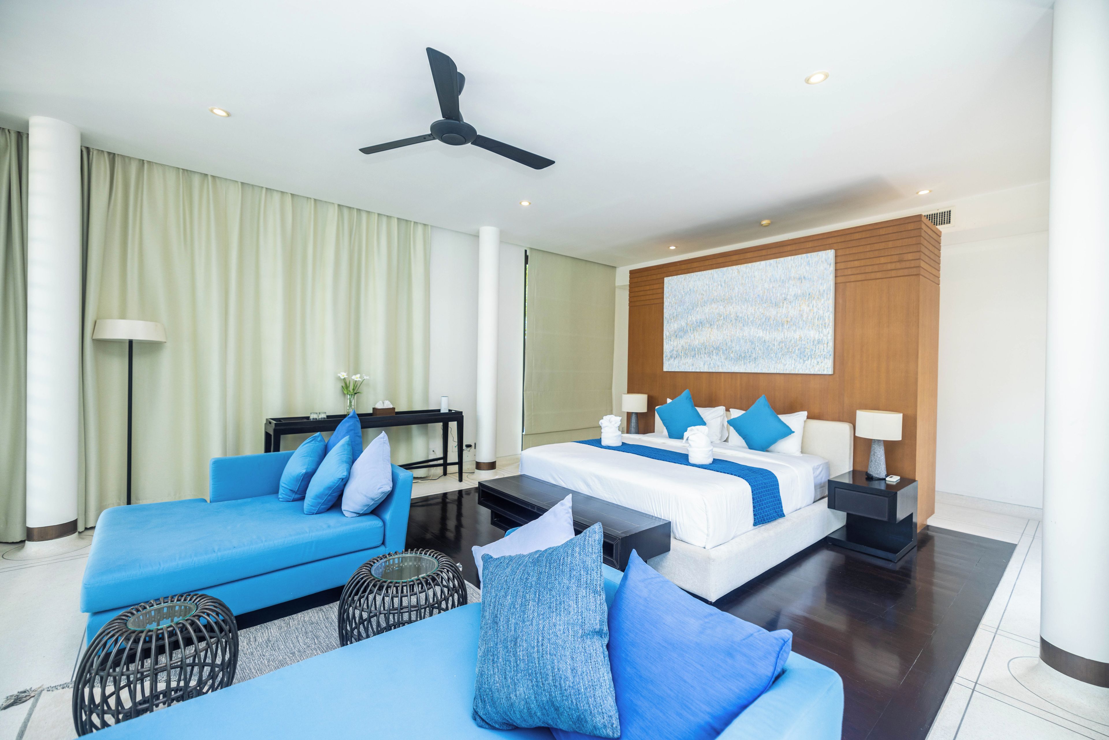 A hotel room with a bed, two blue sofas, a ceiling fan, a desk, and a large painting on the wall.