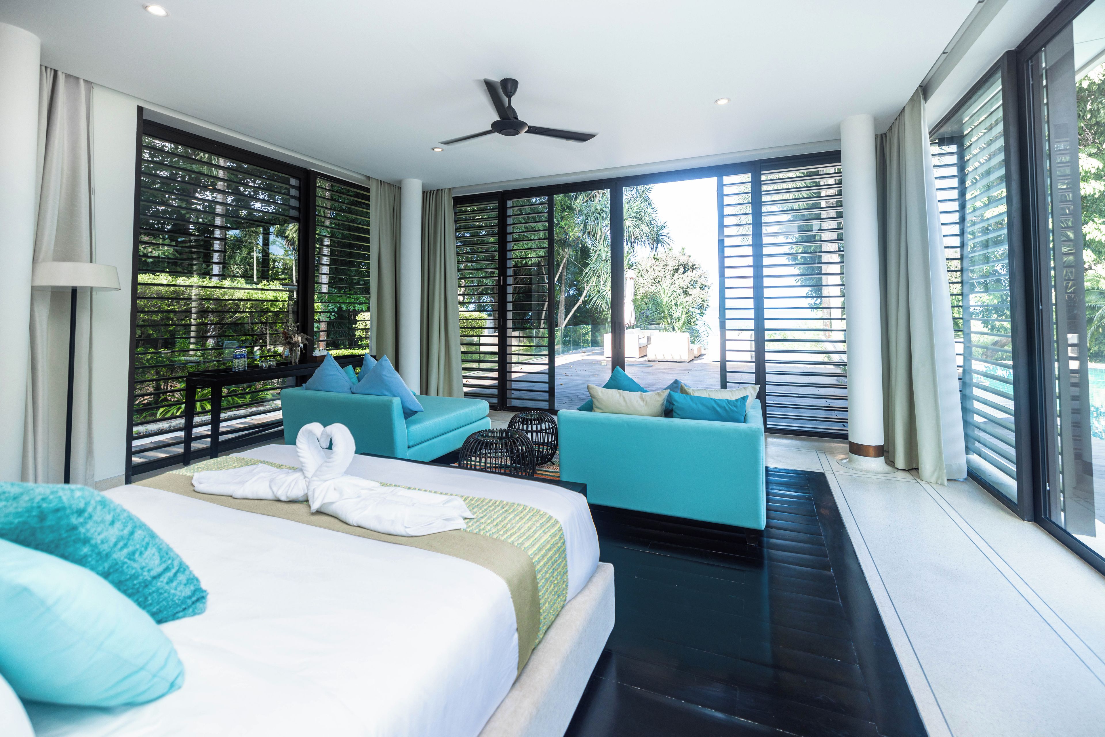Una habitación de hotel moderna con una cama grande, dos sofás, un ventilador de techo y ventanas amplias con persianas.