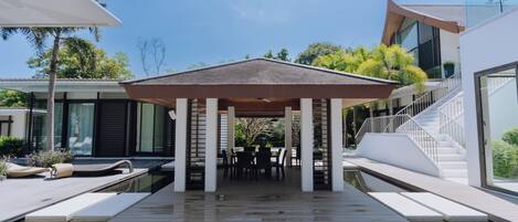 A covered outdoor area with a pool, seating, and a staircase leading to a building.