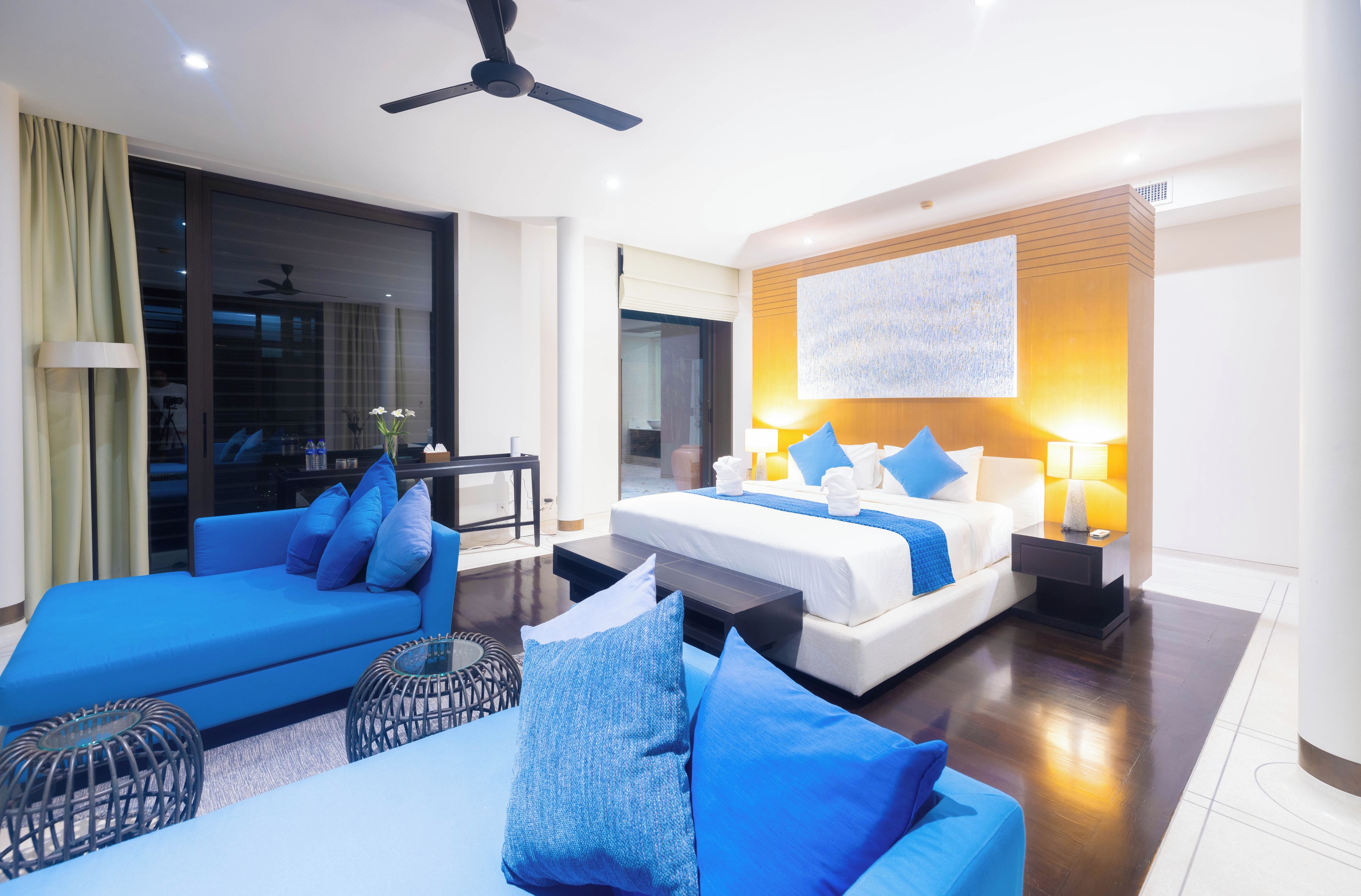 A hotel room with a bed, a sofa, a small table, and a ceiling fan.