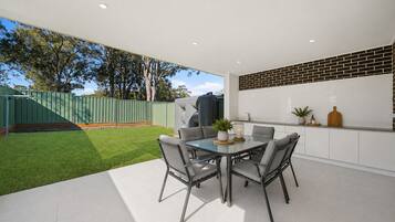 A modern outdoor dining area with a glass table and chairs, a green fence, and a brick wall.