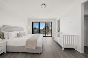 A modern bedroom with a large bed, a white crib, and a balcony with a view.