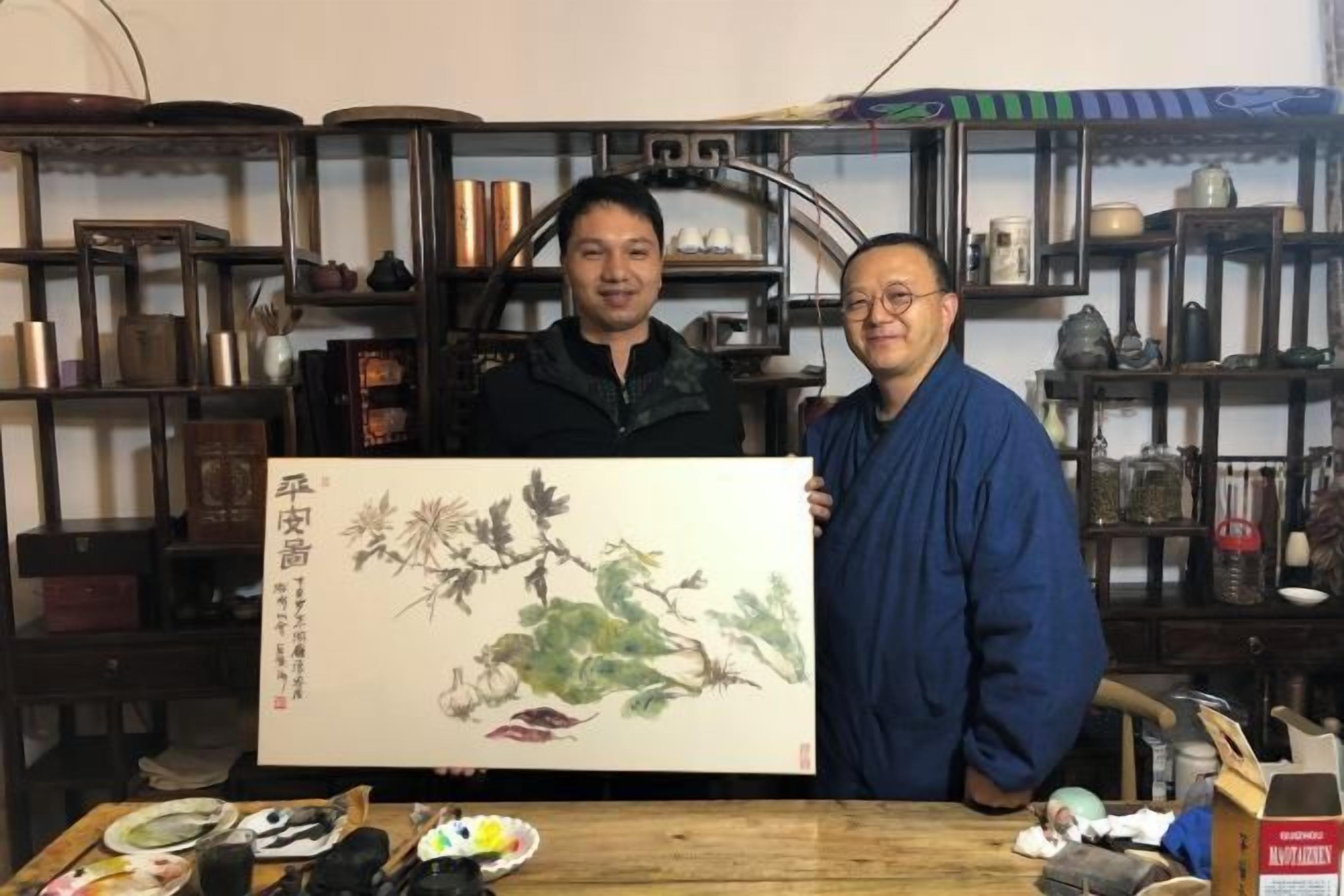 Two individuals holding a large painting in a room with shelves containing various items.