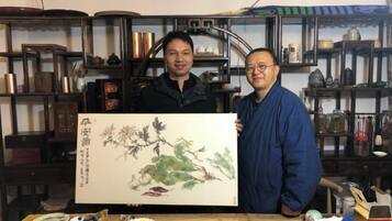 Two individuals holding a large painting in a room with shelves containing various items.