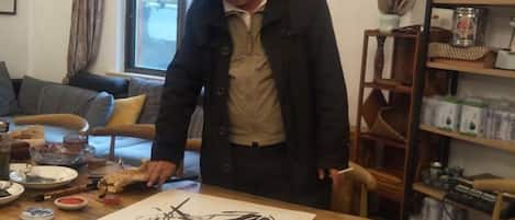 A person standing at a wooden table with a drawing, surrounded by various items and furniture.