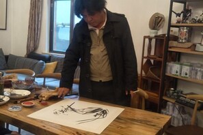 Una persona de pie junto a una mesa de madera con un dibujo, rodeada de varios objetos y muebles.