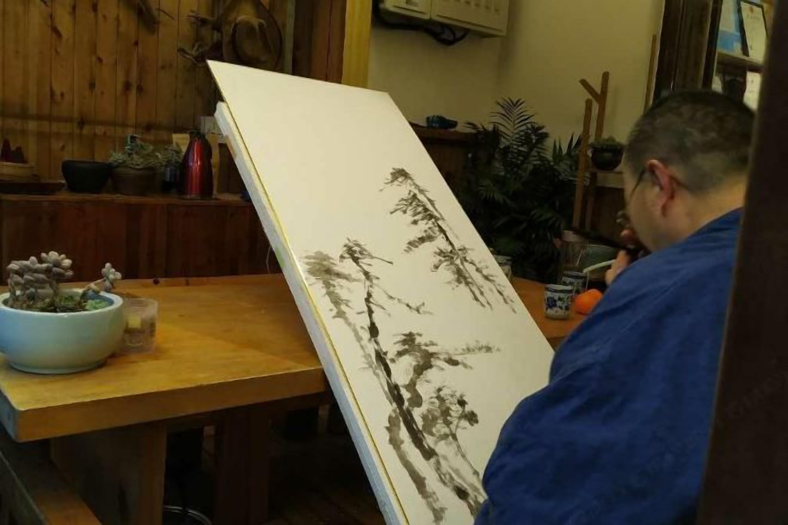 A person in a blue robe standing next to a table with a large white painting and a bowl of flowers.
