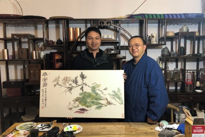 Two individuals holding a large painting in a room with shelves containing various items.