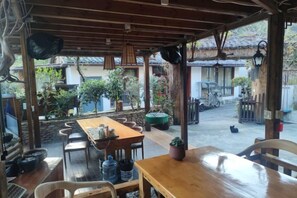 An outdoor dining area with wooden tables and chairs, surrounded by potted plants and a view of a traditional building.