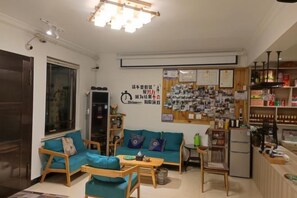 A living room with a sofa, chairs, a coffee table, and a refrigerator. There is a wall with various items and a bulletin board.