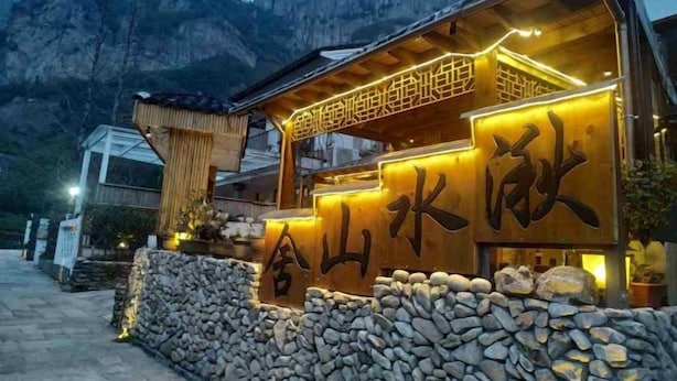 Un edificio con grandes caracteres chinos en la fachada, un muro de piedra al frente y montañas al fondo.