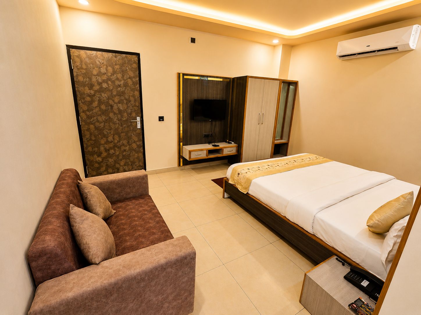 A hotel room with a bed, a sofa, a TV, and an air conditioning unit.