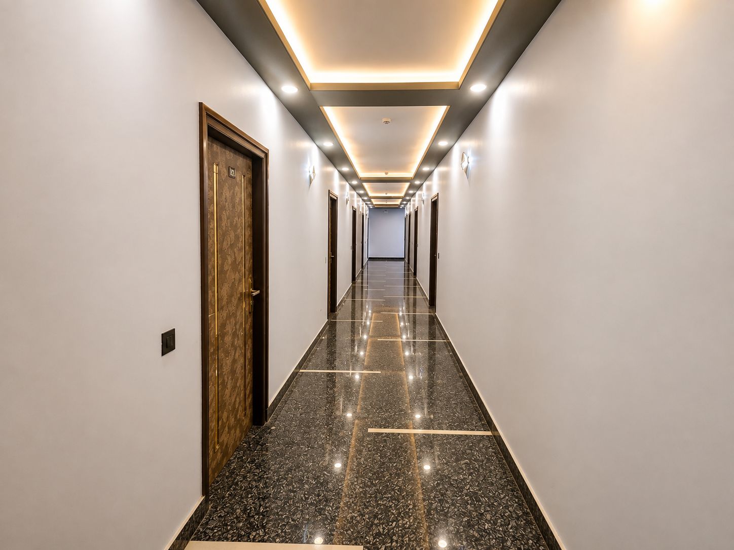Un largo pasillo bien iluminado con piso pulido, puertas a ambos lados e iluminación empotrada en el techo.