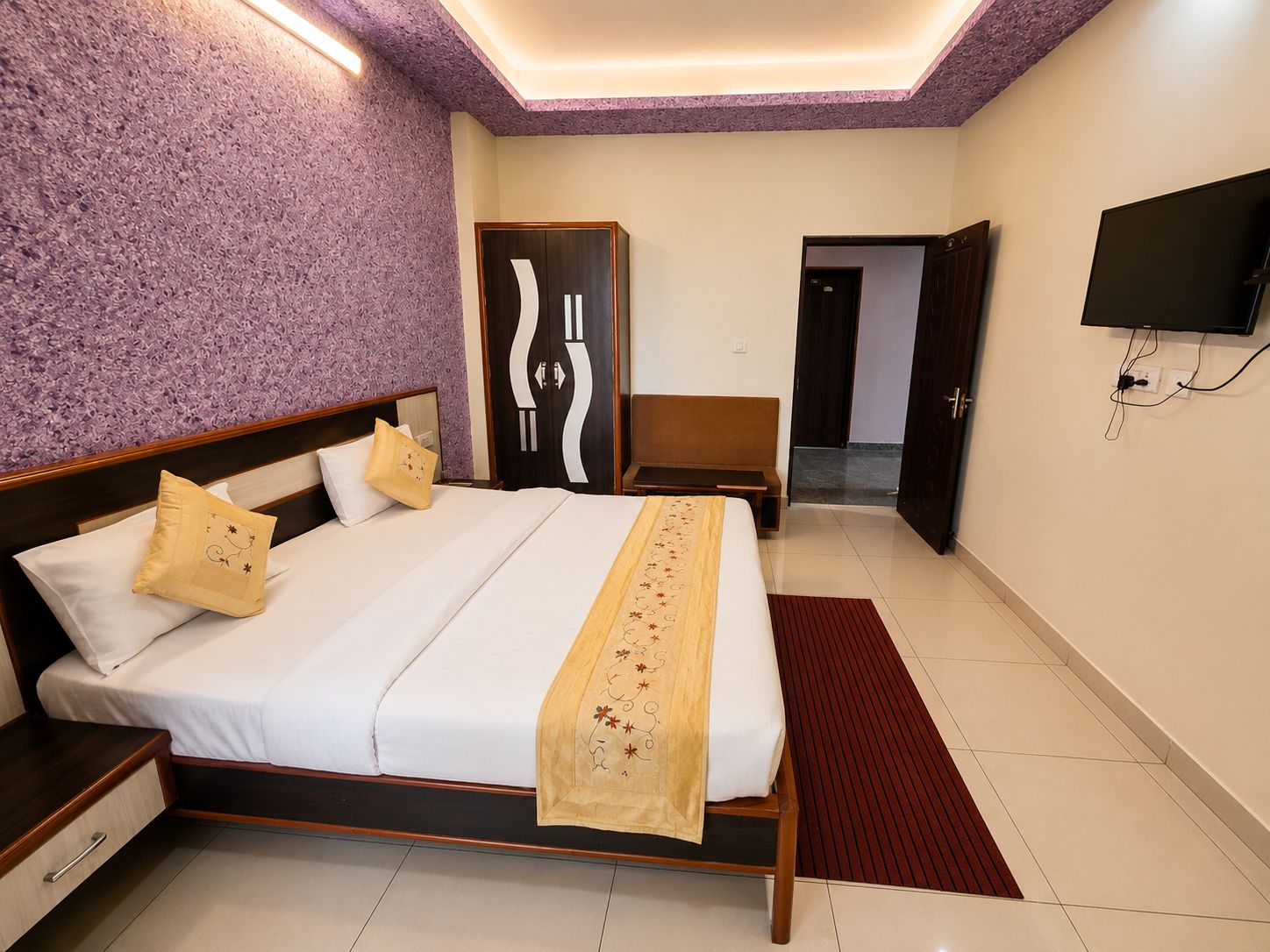 A hotel room with a bed, a wooden nightstand, a television mounted on the wall, and a door leading to another room.