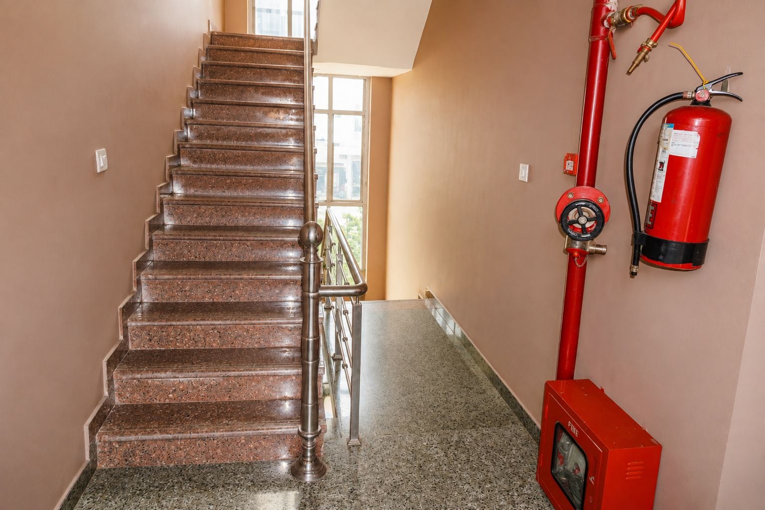 Uma escada com corrimão de metal, um extintor fixado na parede e uma mangueira de incêndio.