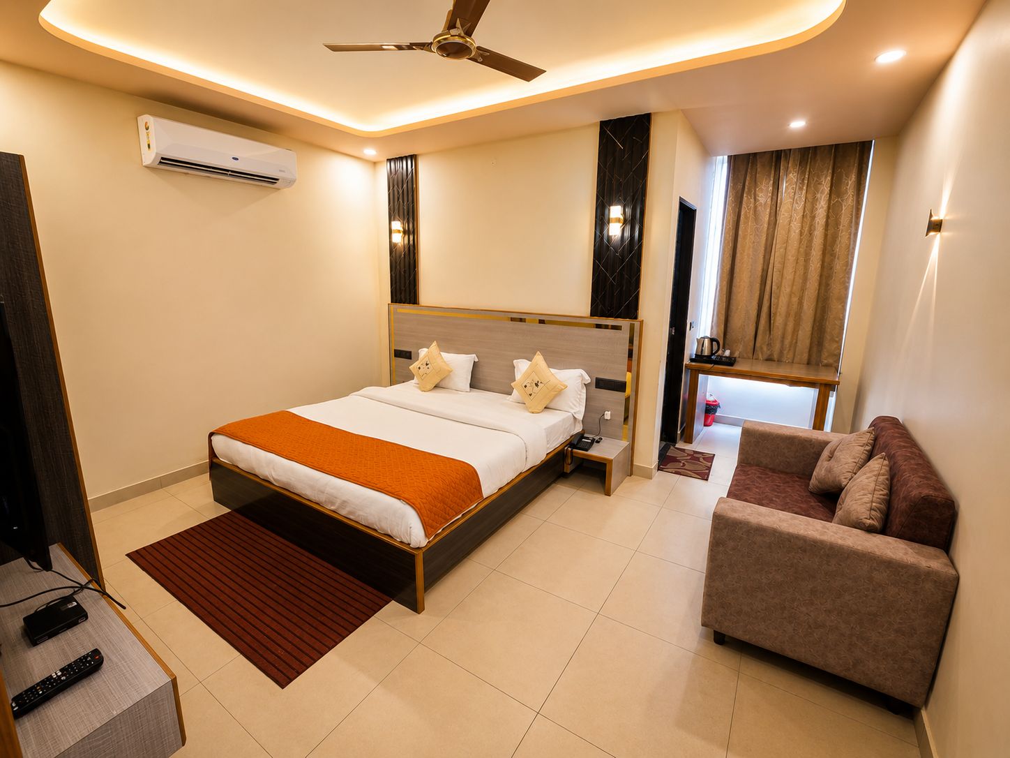 Habitación de hotel con cama, sofá, ventilador de techo, aire acondicionado y ventana con cortinas.