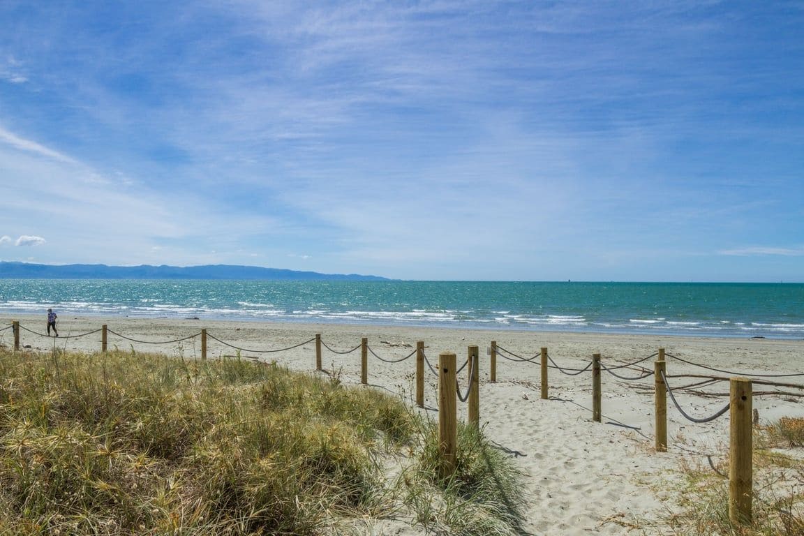 A beach with a wooden fence leading to the shore, a clear sky, and a person walking on the sand.