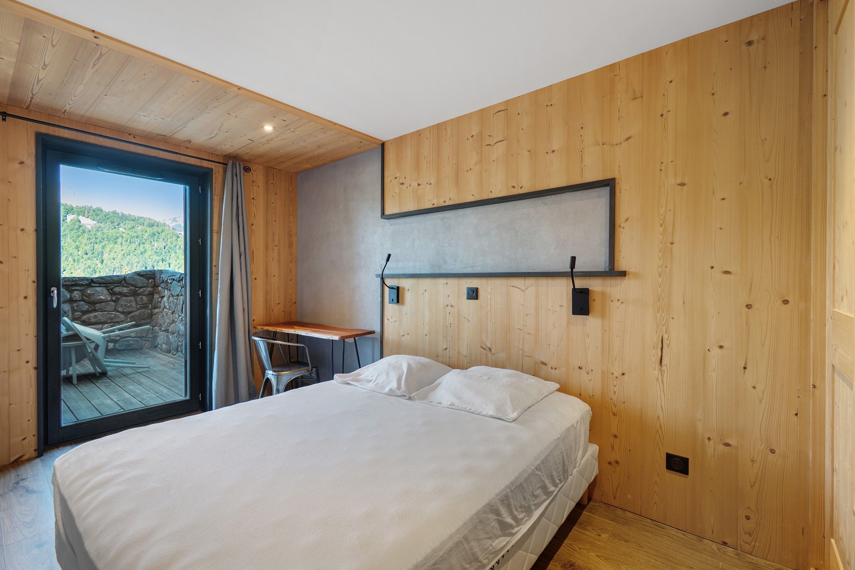 Une chambre à coucher avec un lit, un bureau et une vue sur un paysage montagneux, visible à travers une porte vitrée.