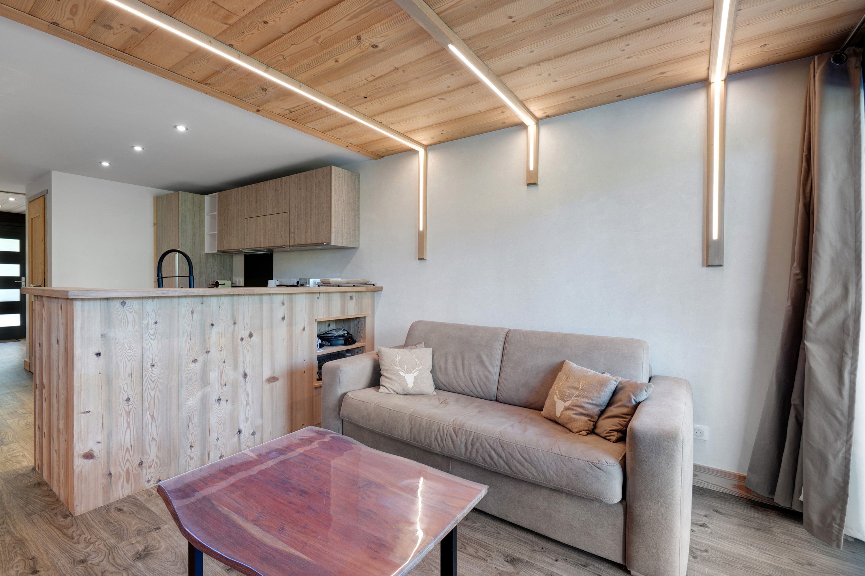 Un intérieur moderne avec un plafond en bois, un coin cuisine, un canap é agrémenté de coussins décoratifs et une table basse en verre.