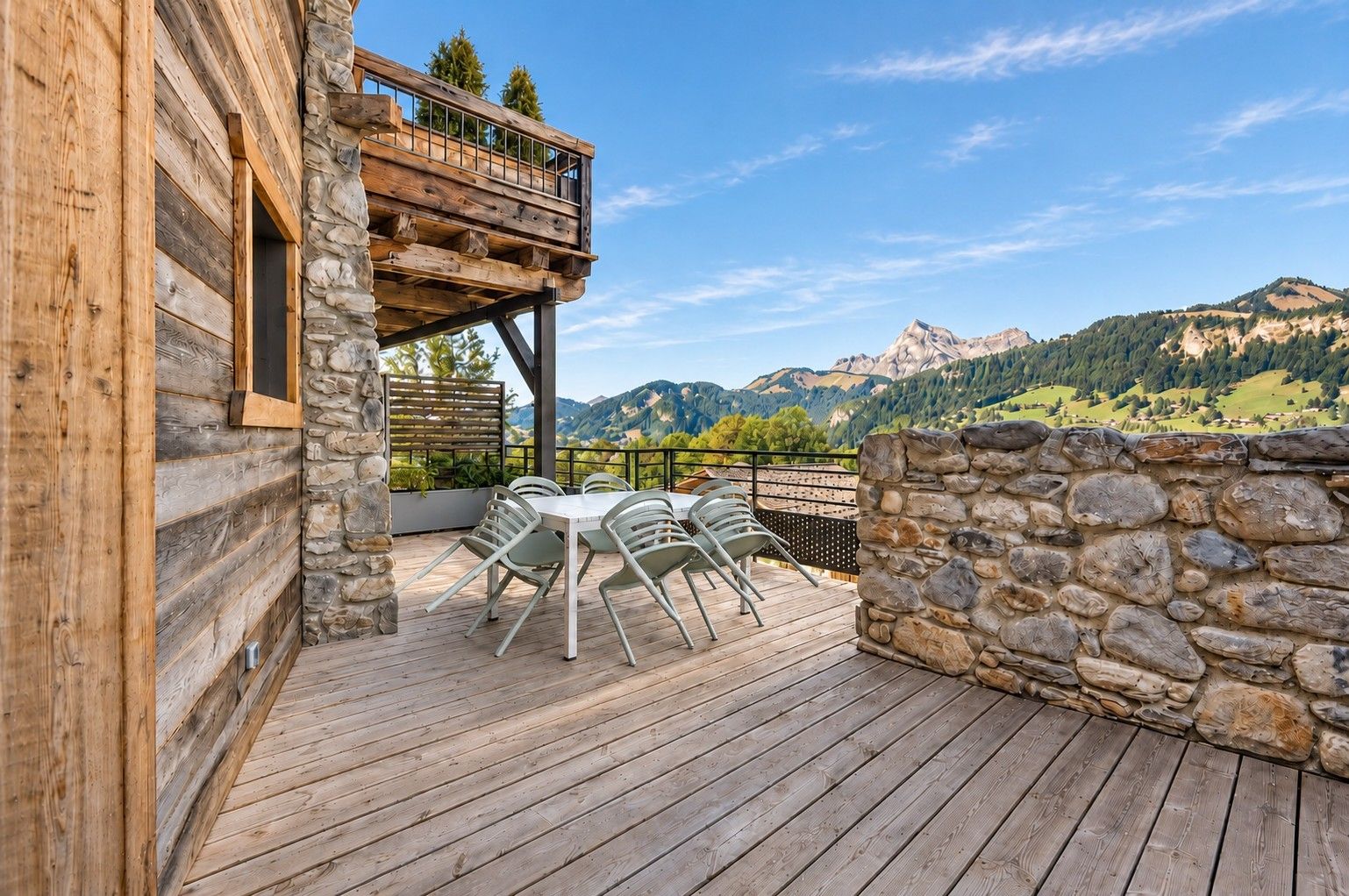 Une terrasse en bois, agrémentée de mobilier d’extérieur et bordée d’un mur en pierre, qui offre une vue sur un paysage montagneux.