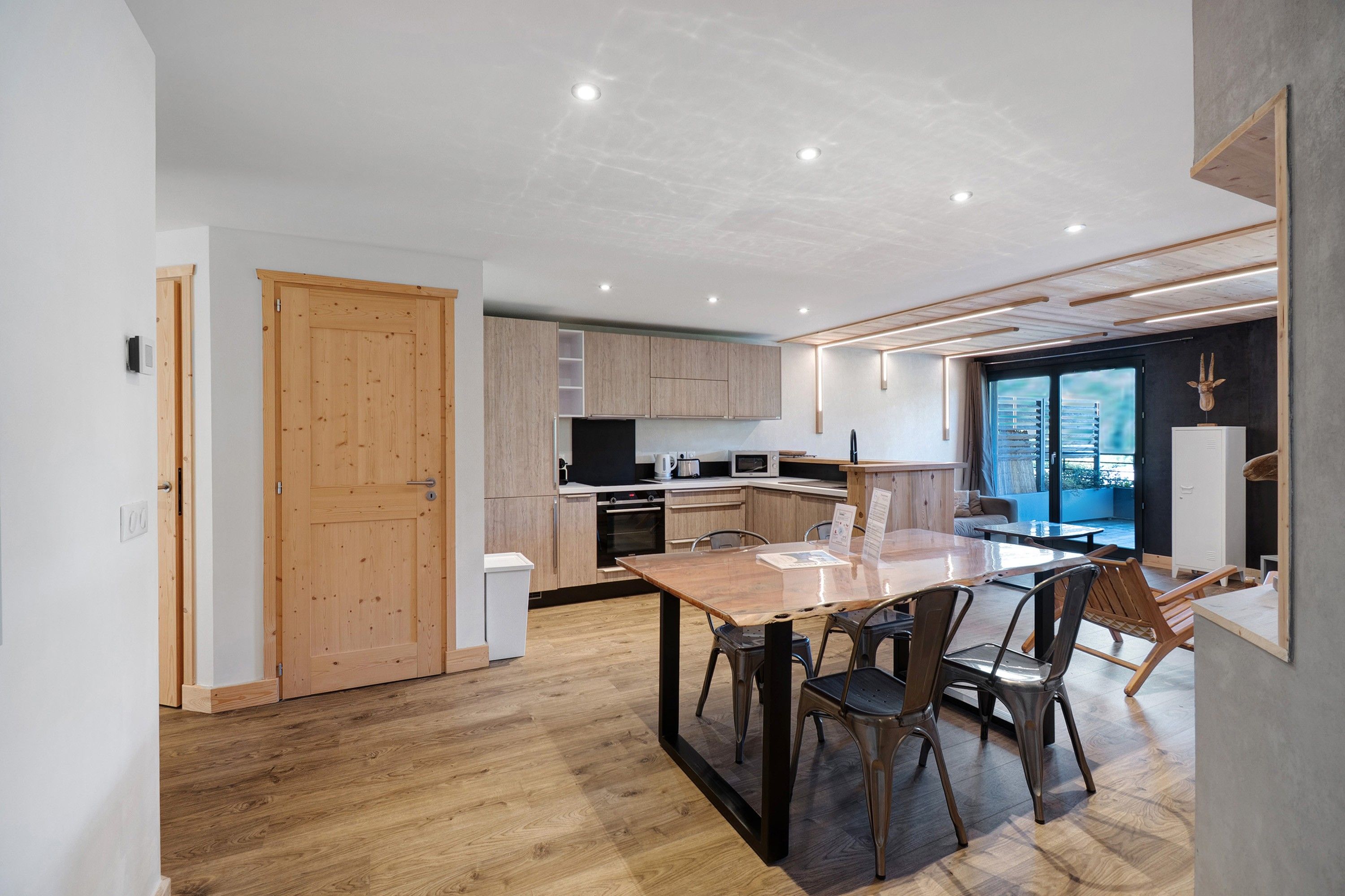 Une cuisine moderne avec des meubles en bois, une table à manger avec des chaises et une vue sur l’extérieur grâce à une grande fenêtre.