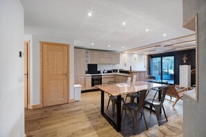 Eine moderne Küche mit Holzausstattung, einem Esstisch mit Stühlen und Blick ins Freie durch ein großes Fenster.