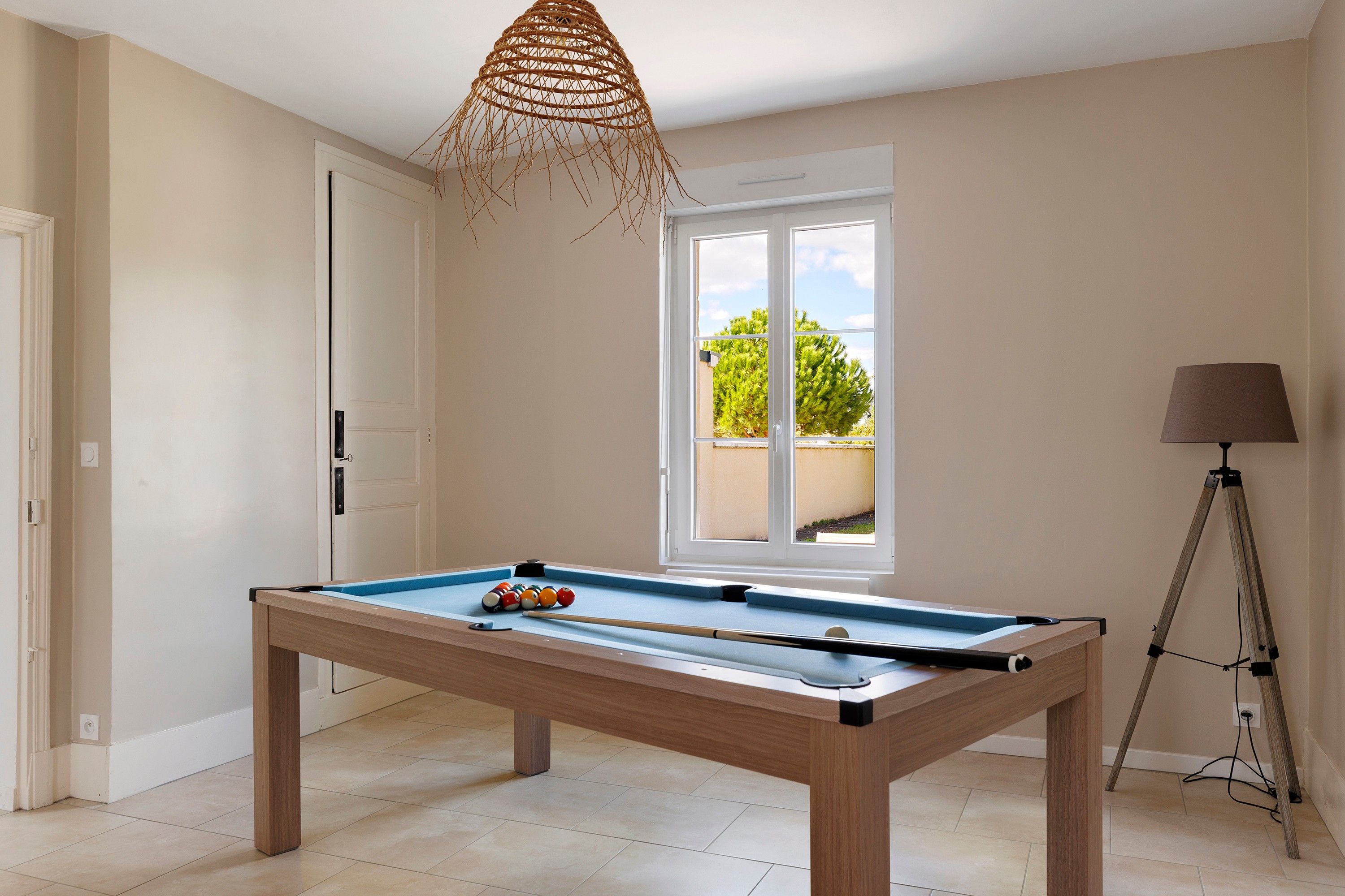 A pool table in a room with a door, a window with a view of greenery, and a tripod lamp.
