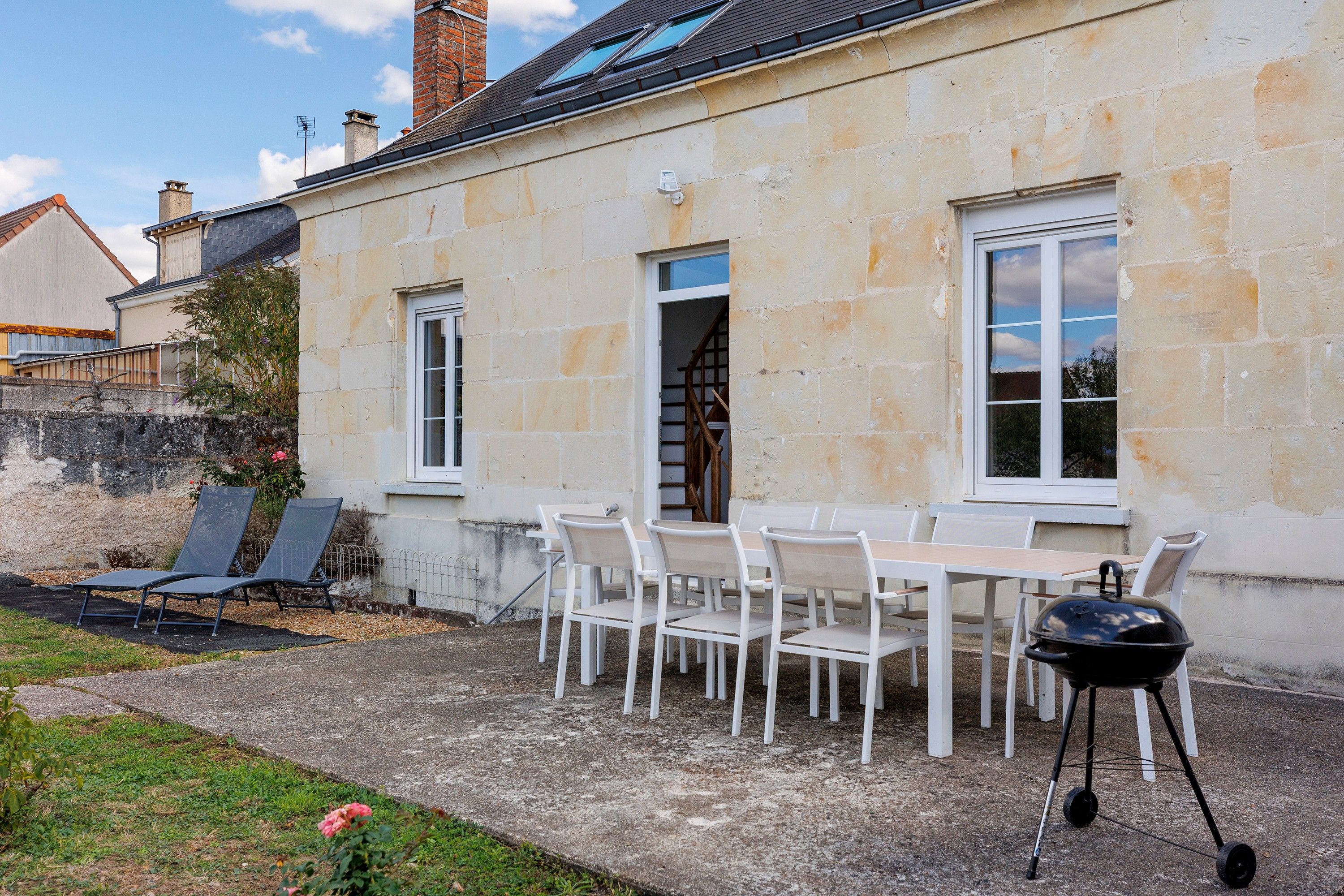 A stone building with a patio area featuring a table and chairs, a barbecue grill, and a garden with a rose bush.