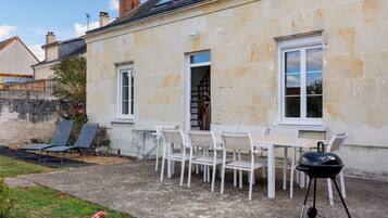 A stone building with a patio area featuring a table and chairs, a barbecue grill, and a garden with a rose bush.