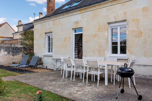 Ein Steinhaus mit einer Terrassenanlage, komplett mit Tisch, Stühlen und einem Grill, sowie ein Garten mit einem Rosenbusch.
