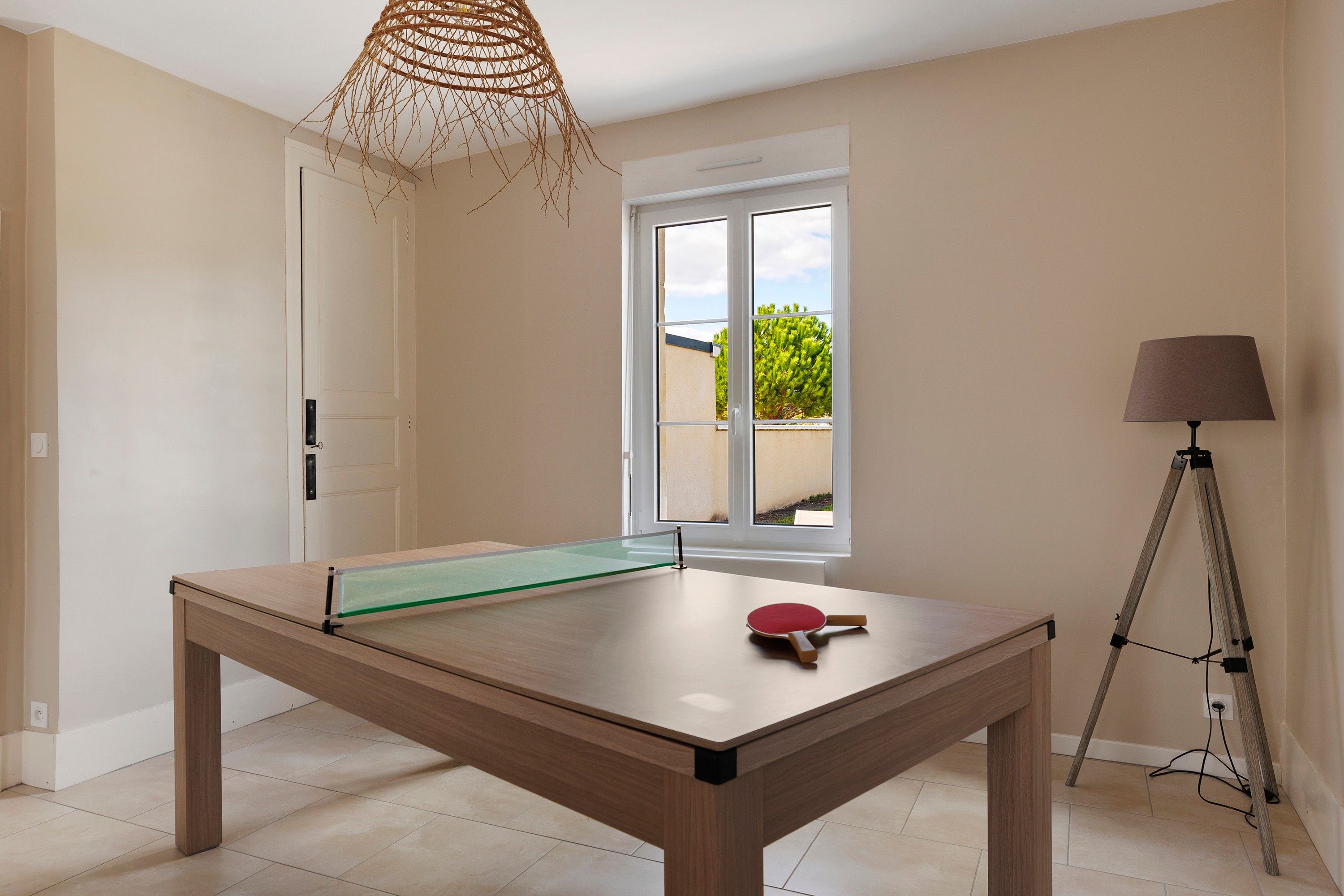 A ping pong table in a room with a door, a window with a view of greenery, and a tripod lamp.