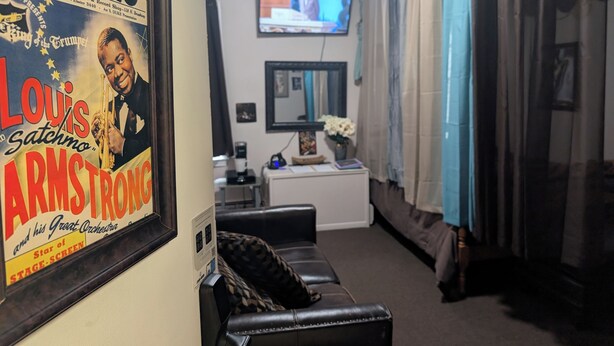 A living room with a framed poster of Louis Armstrong, a television, a couch, and a coffee table. - 146-2 Satchmo, Louie Armstrong. Chocolatey Jazz Lounge w/Mini Rolling Desk. (New London)