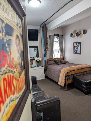 A bedroom with a bed, a bedside table, a mirror, a wall-mounted artwork, and a poster. - 146-2 Satchmo, Louie Armstrong. Chocolatey Jazz Lounge w/Mini Rolling Desk. (New London)