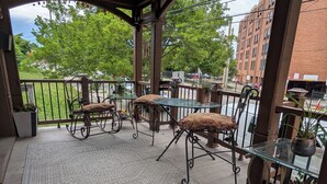 A covered outdoor seating area with metal chairs and tables, surrounded by potted plants and a view of a building and trees. - 146-2 Satchmo, Louie Armstrong. Chocolatey Jazz Lounge w/Mini Rolling Desk. (New London)