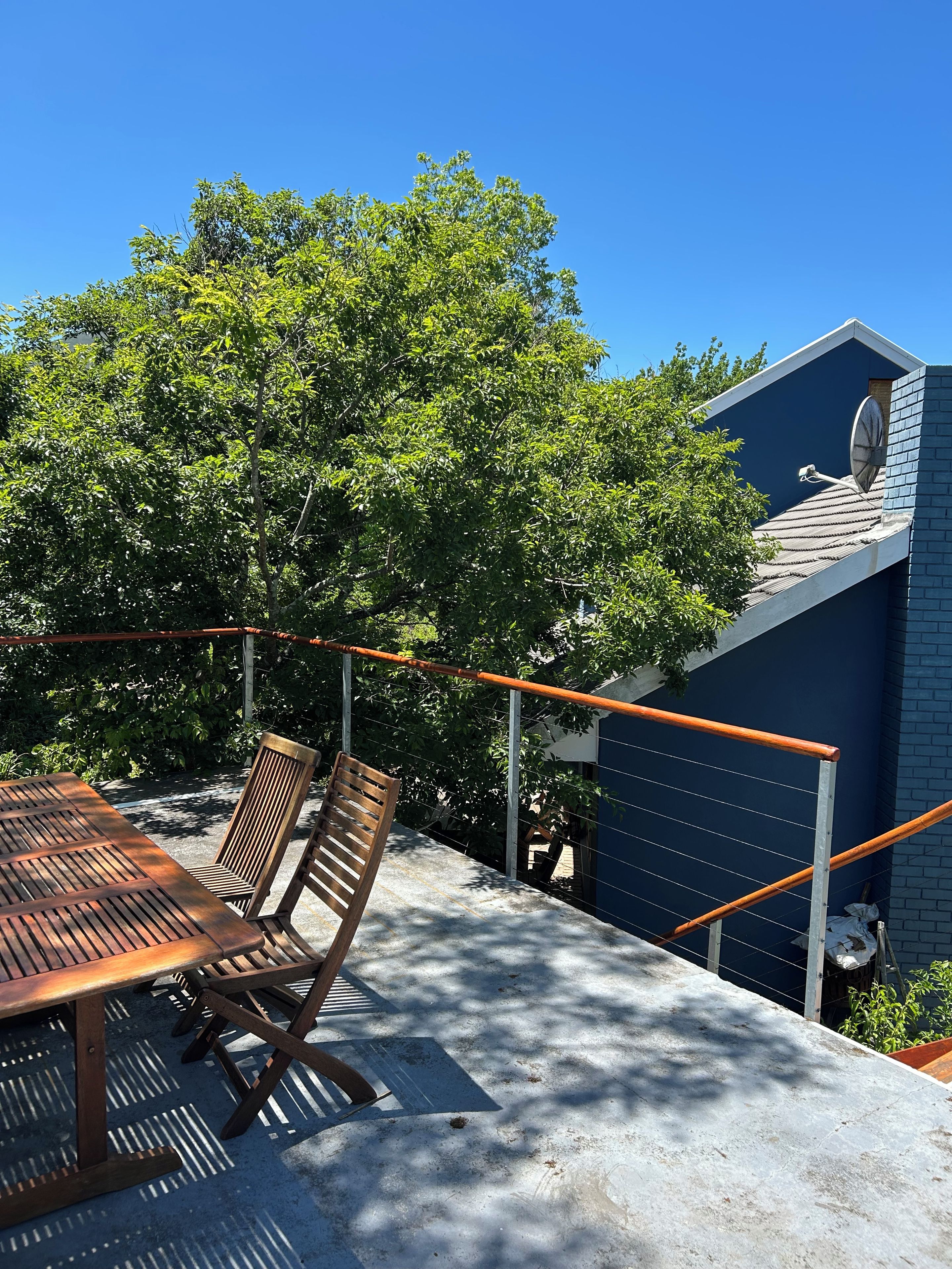 An outdoor seating area with a table and chairs, a metal railing, and a tree providing shade.